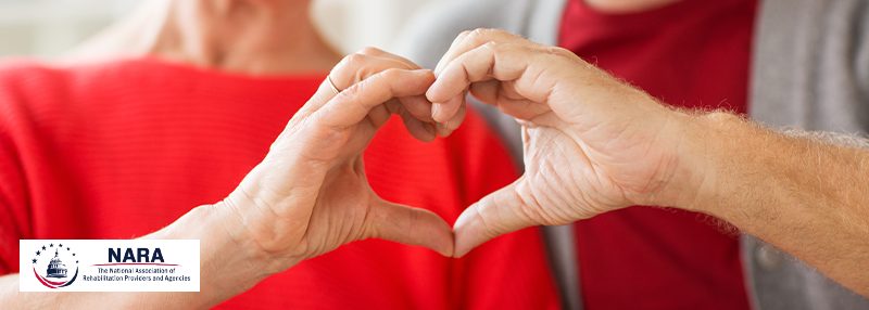 A middle-aged couple wearing red shirts using their hands to make a heart