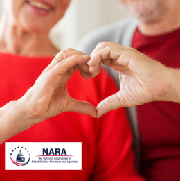 A middle-aged couple wearing red shirts using their hands to make a heart